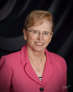 JUDY AUG_2012 - Serra Club of Philadelphia Smiling older woman with short light hair and glasses, wearing a pink blazer against a dark backdrop.